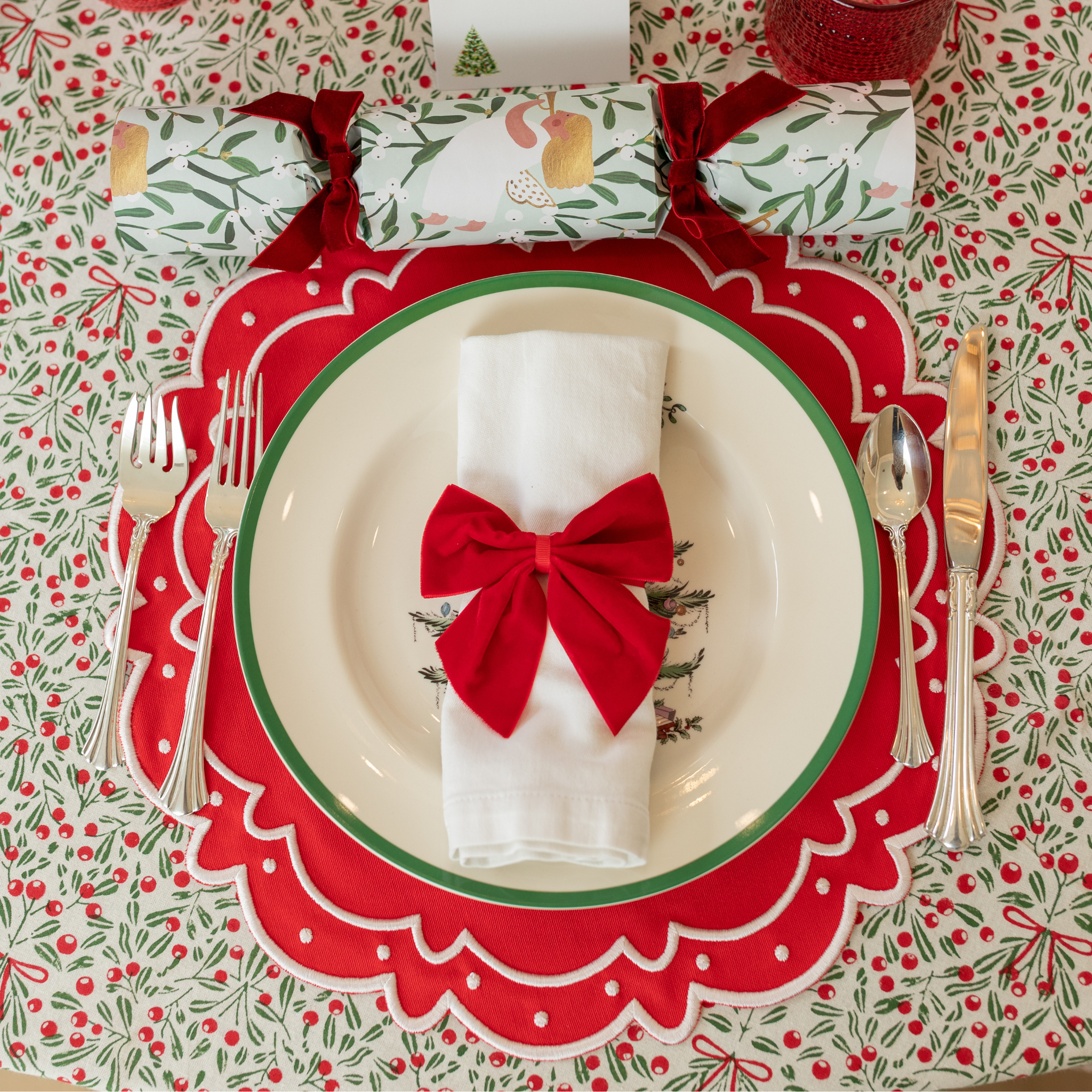 Red velvet bow napkin ties on white napkins, styled on a festive holiday table with holly tablecloth and scalloped placemats – perfect for Christmas entertaining and seasonal celebrations.