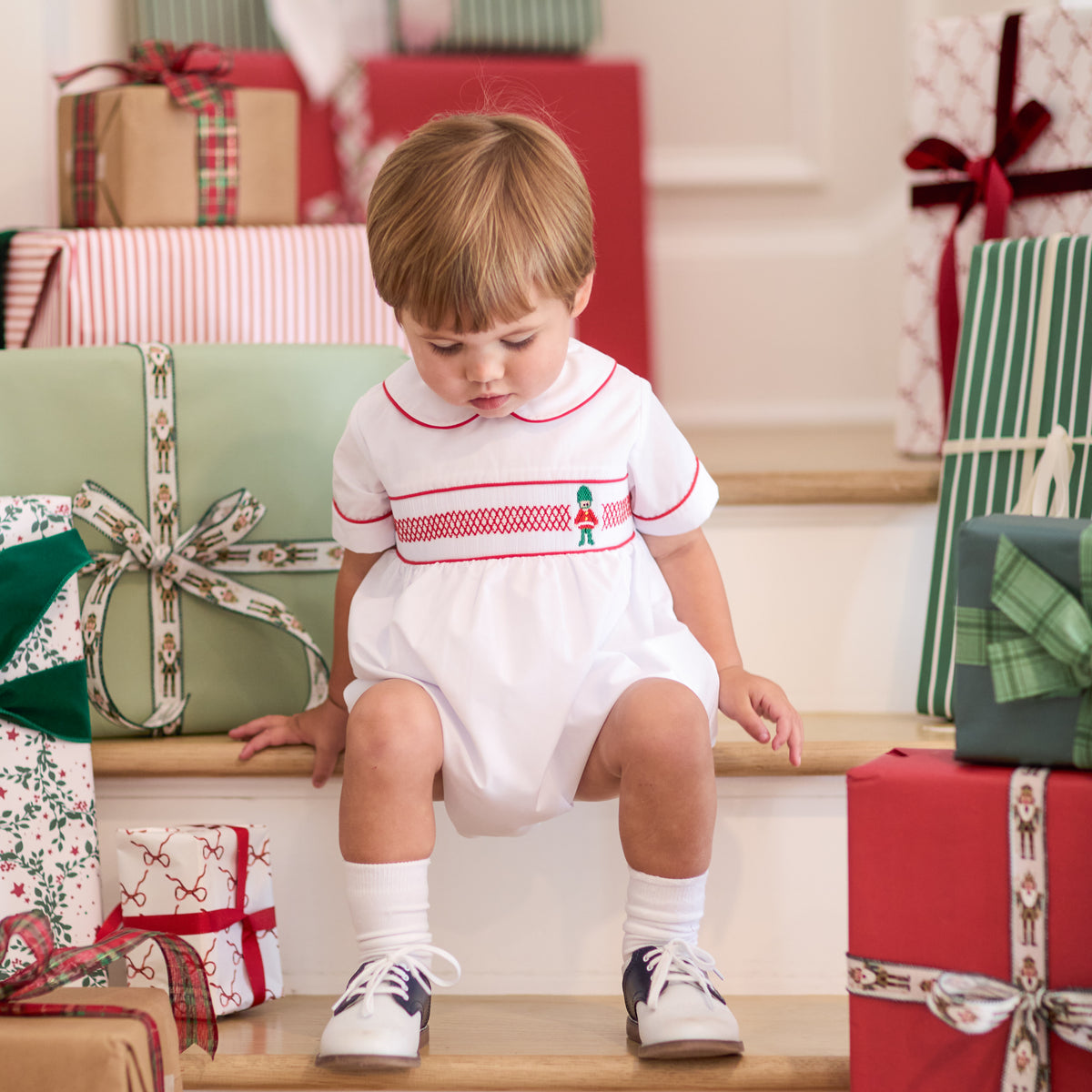 White baby boy holiday bubble with red toy soldier smocking and Peter Pan collar, Henry Duvall's Charlie Bubble in cotton poplin for Christmas celebrations