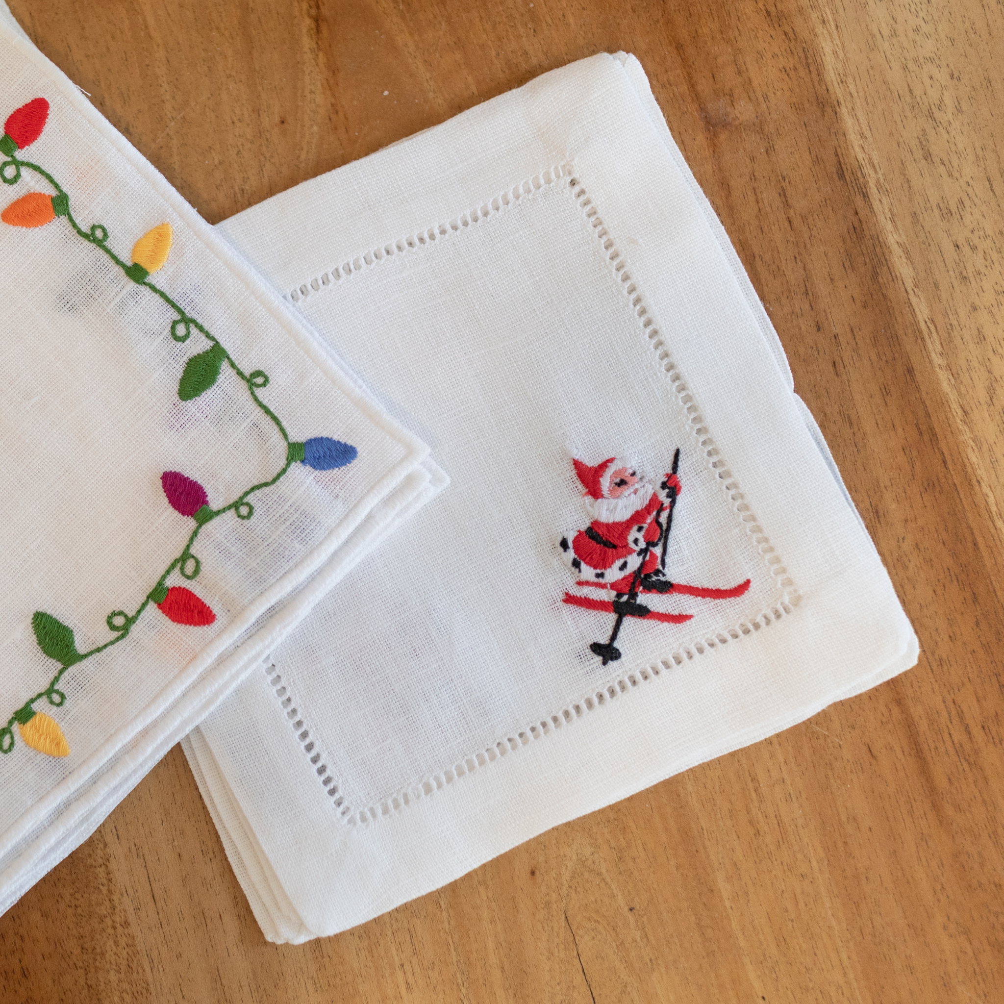 Four white cotton cocktail napkins featuring a red and black embroidered skiing Santa design.