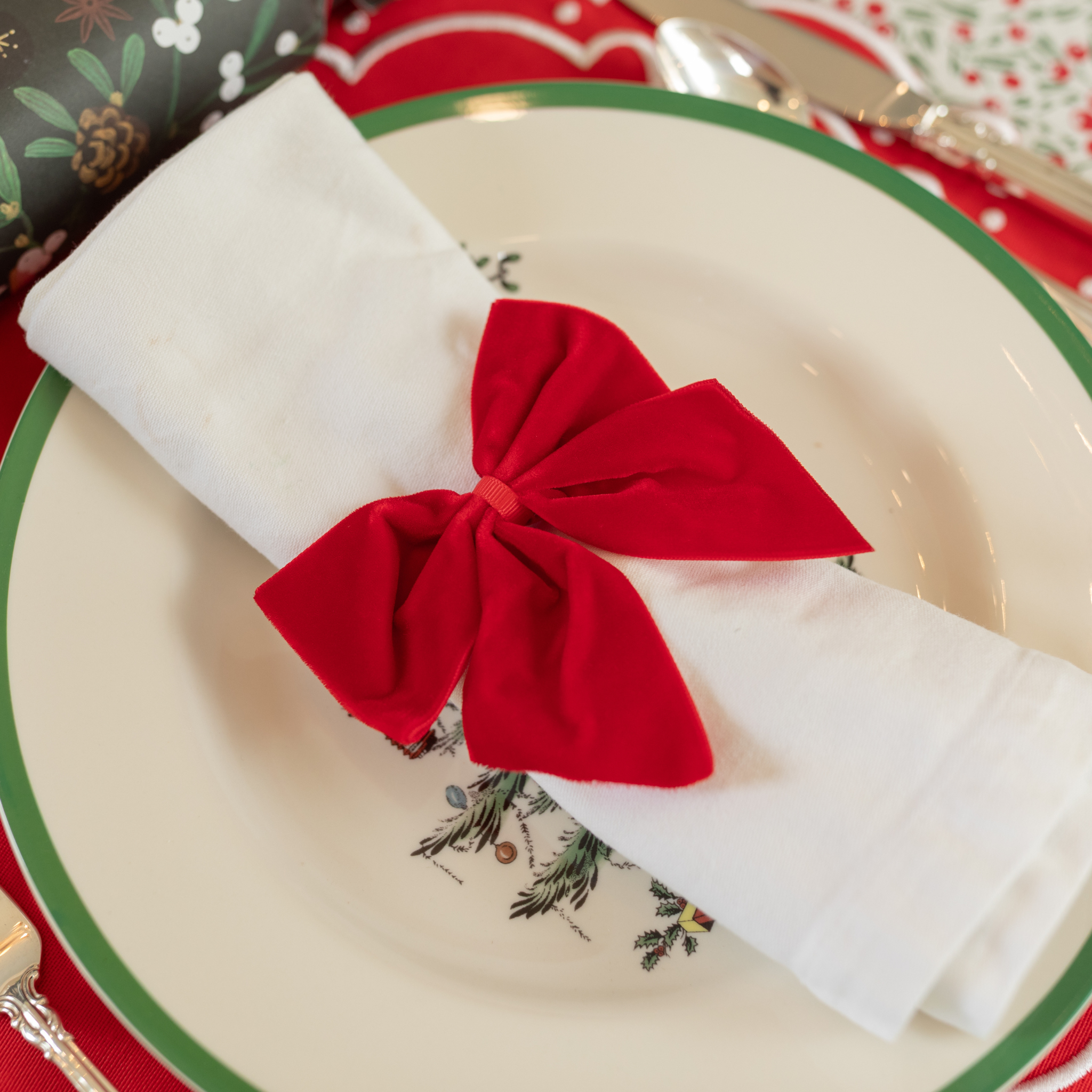Red velvet bow napkin ties on white napkins, styled on a festive holiday table with holly tablecloth and scalloped placemats – perfect for Christmas entertaining and seasonal celebrations.