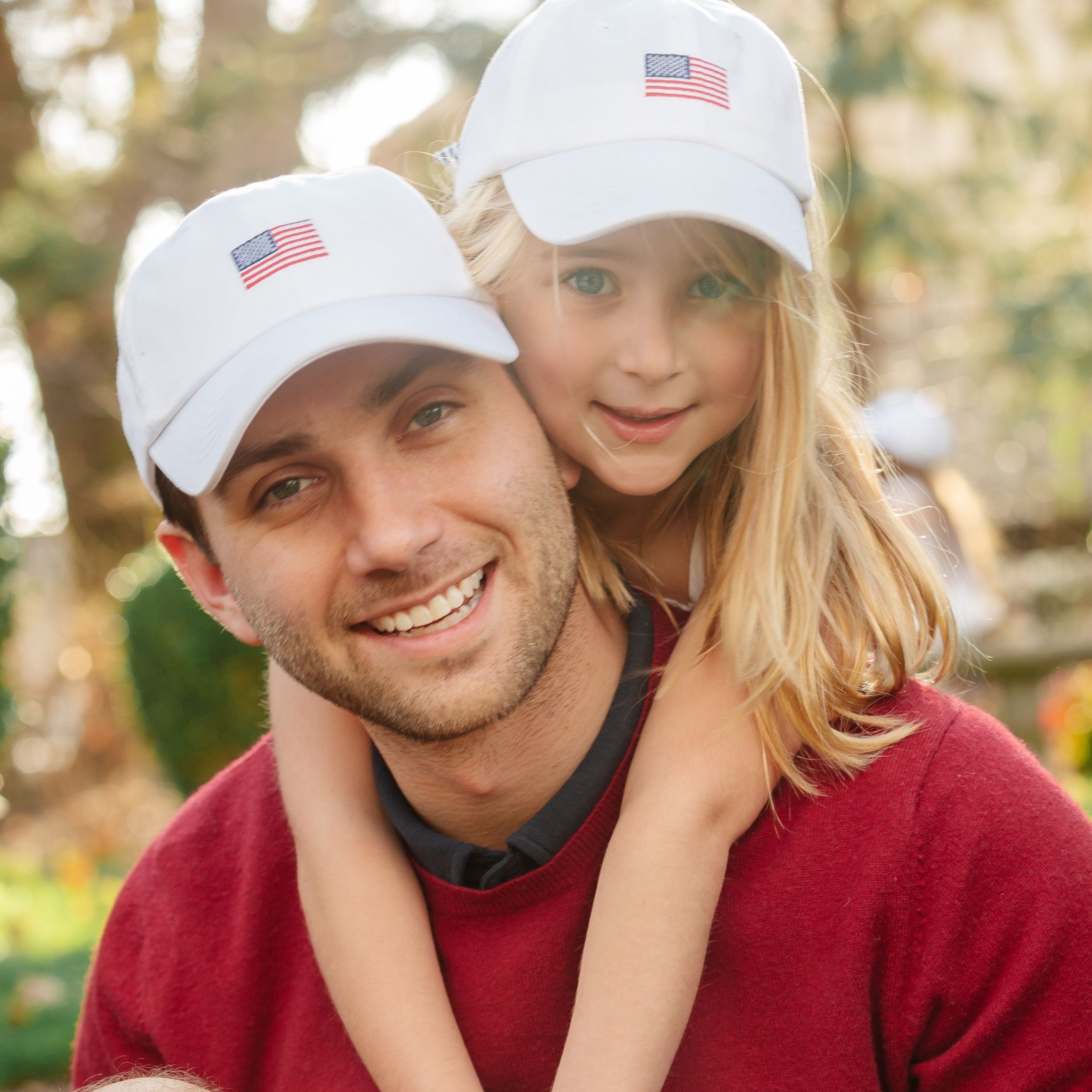Man holding a young girl on his shoulders, both wearing white hats with American flags.