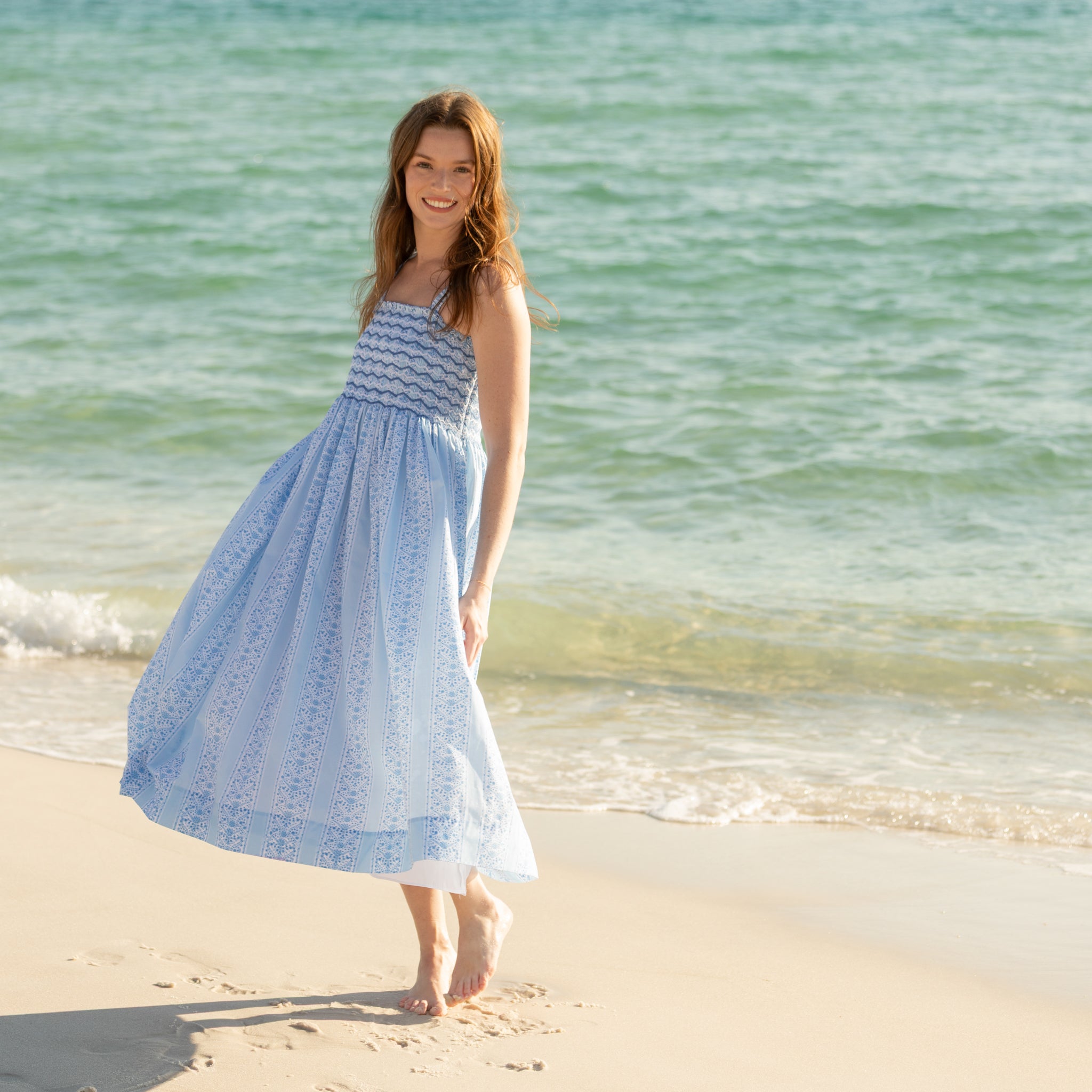 Woman walking along the beach wearing the Henry Duvall Kate Dress, a breezy striped women’s dress with coastal style.