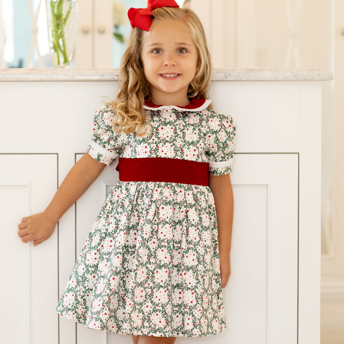 Girls’ holiday dress in Henry Duvall Regent Wreaths red and green Christmas print, featuring a red velvet Peter Pan collar and sash, white pleated trim, puff sleeves, and a full gathered skirt.
