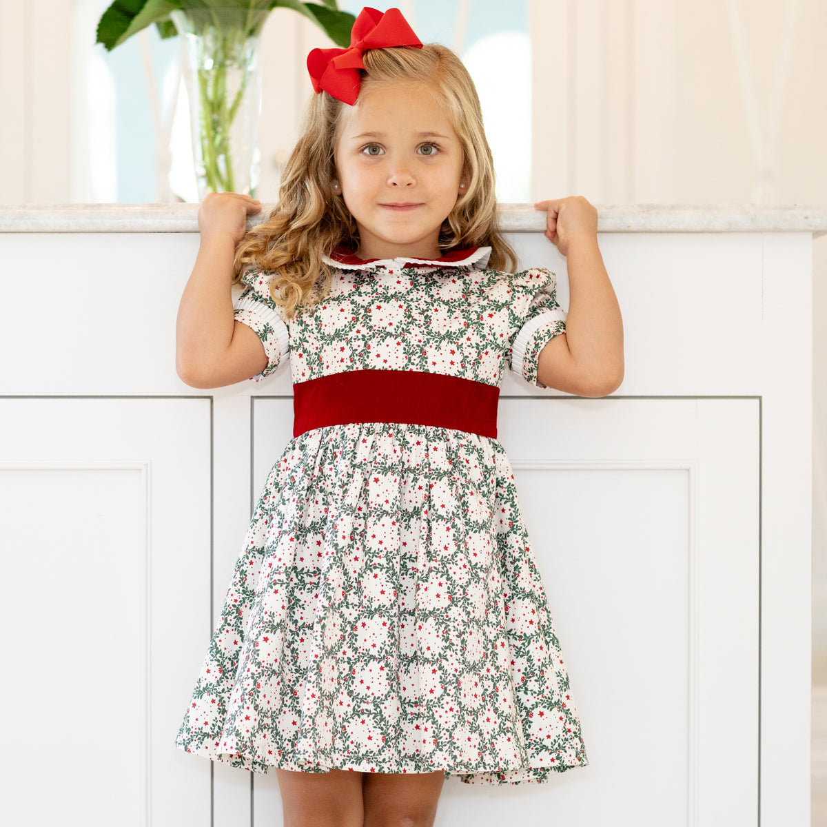 Girls’ holiday dress in Henry Duvall Regent Wreaths red and green Christmas print, featuring a red velvet Peter Pan collar and sash, white pleated trim, puff sleeves, and a full gathered skirt.