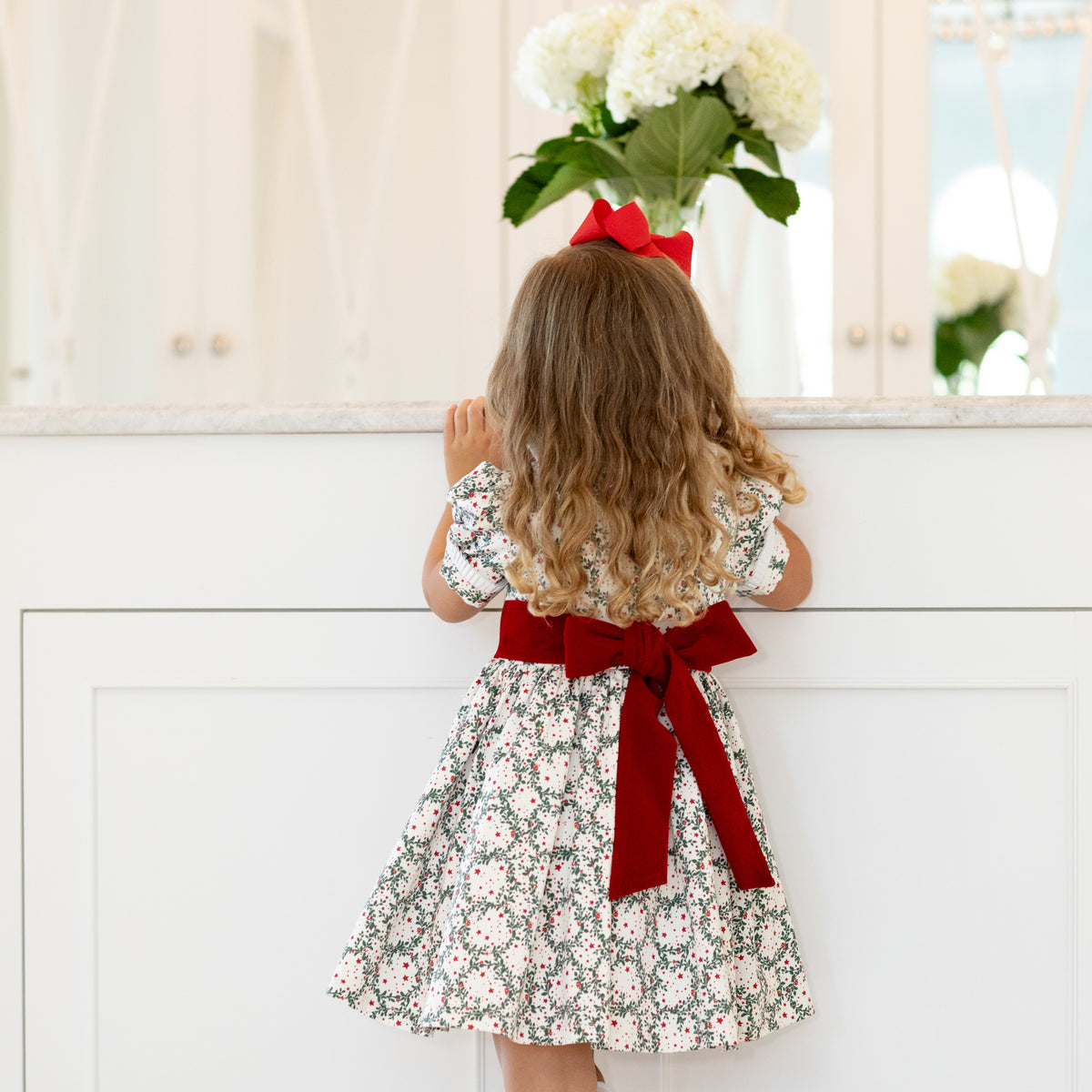 Girls’ holiday dress in Henry Duvall Regent Wreaths red and green Christmas print, featuring a red velvet Peter Pan collar and sash, white pleated trim, puff sleeves, and a full gathered skirt.