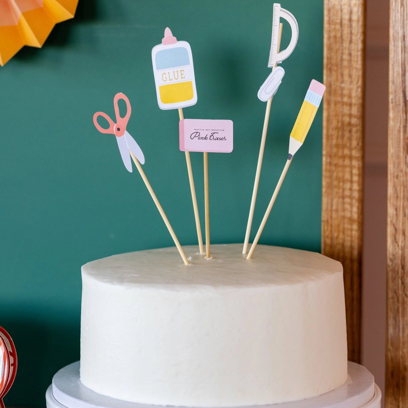 White cake with decorative toppers on a stand against a green wall.