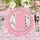 Table setting with pink placemat, white plates, and silverware on a floral tablecloth.