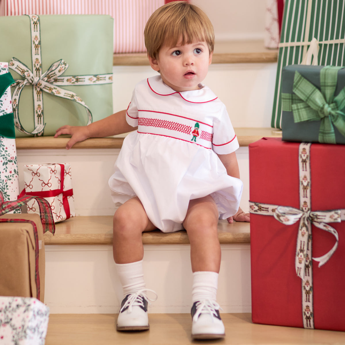 White baby boy holiday bubble with red toy soldier smocking and Peter Pan collar, Henry Duvall's Charlie Bubble in cotton poplin for Christmas celebrations