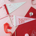 Packaged set of two festive felt pennants with scalloped edges, one in cream with “On the Nice List” in red and pink, and one in soft pink with “On the Naughty List,” ideal for kids' Christmas parties or playful holiday decor