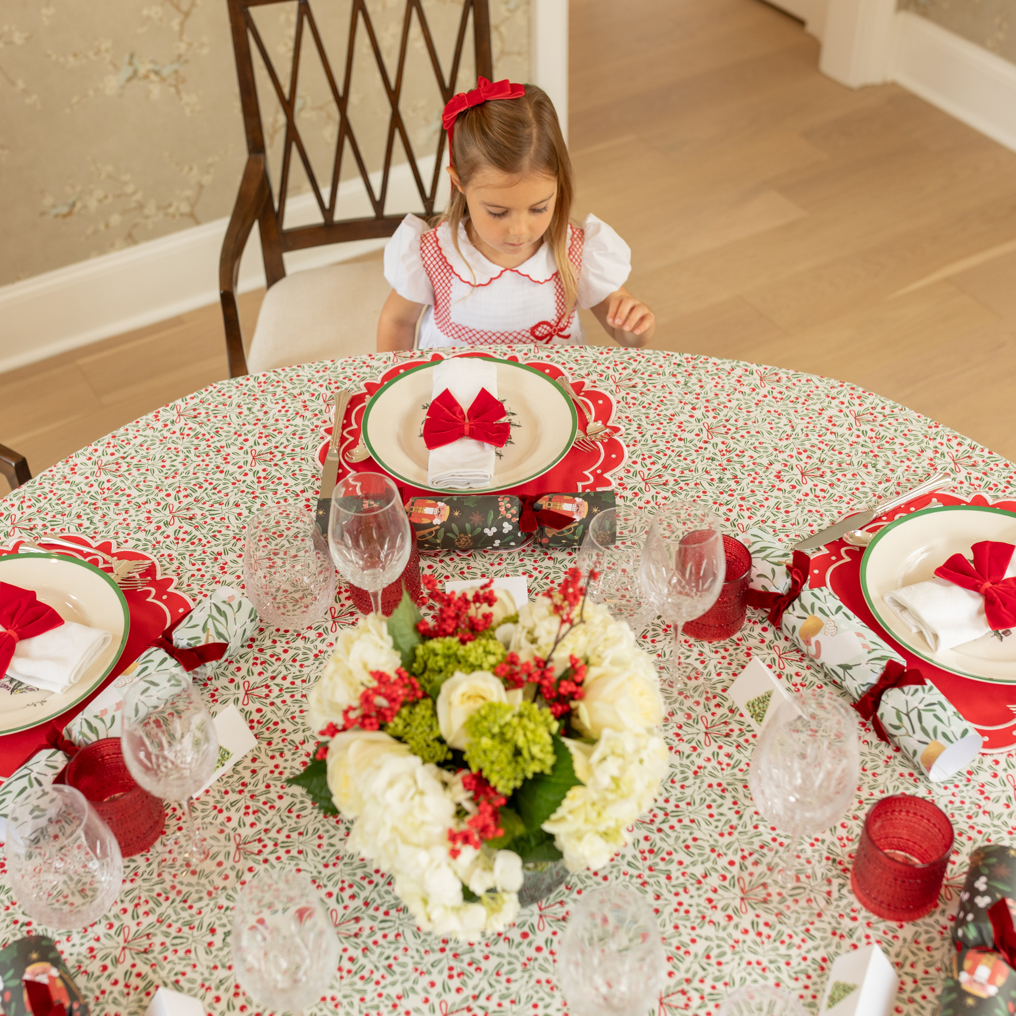 Red velvet bow napkin ties on white napkins, styled on a festive holiday table with holly tablecloth and scalloped placemats – perfect for Christmas entertaining and seasonal celebrations.