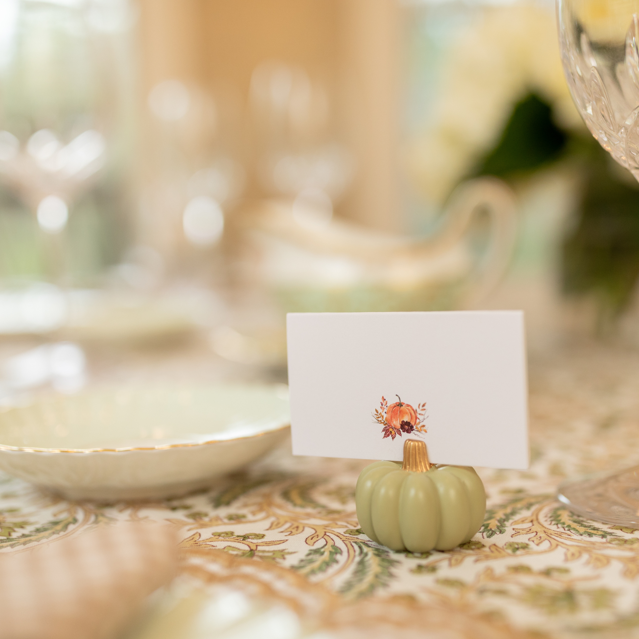 Watercolor Thanksgiving place card featuring a pumpkin illustration, displayed in a pale green pumpkin holder on a patterned fall tablecloth.