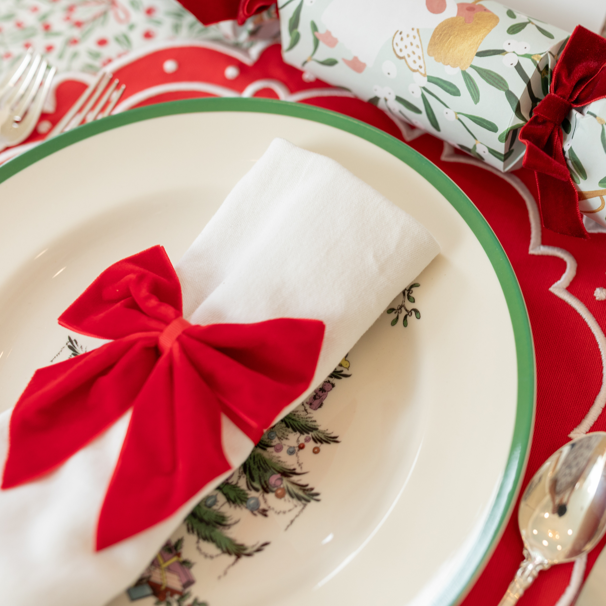 Red velvet bow napkin ties on white napkins, styled on a festive holiday table with holly tablecloth and scalloped placemats – perfect for Christmas entertaining and seasonal celebrations.