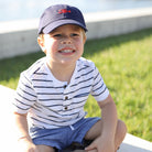 Child wearing a striped shirt and cap sitting on a dock with grass and water in the background