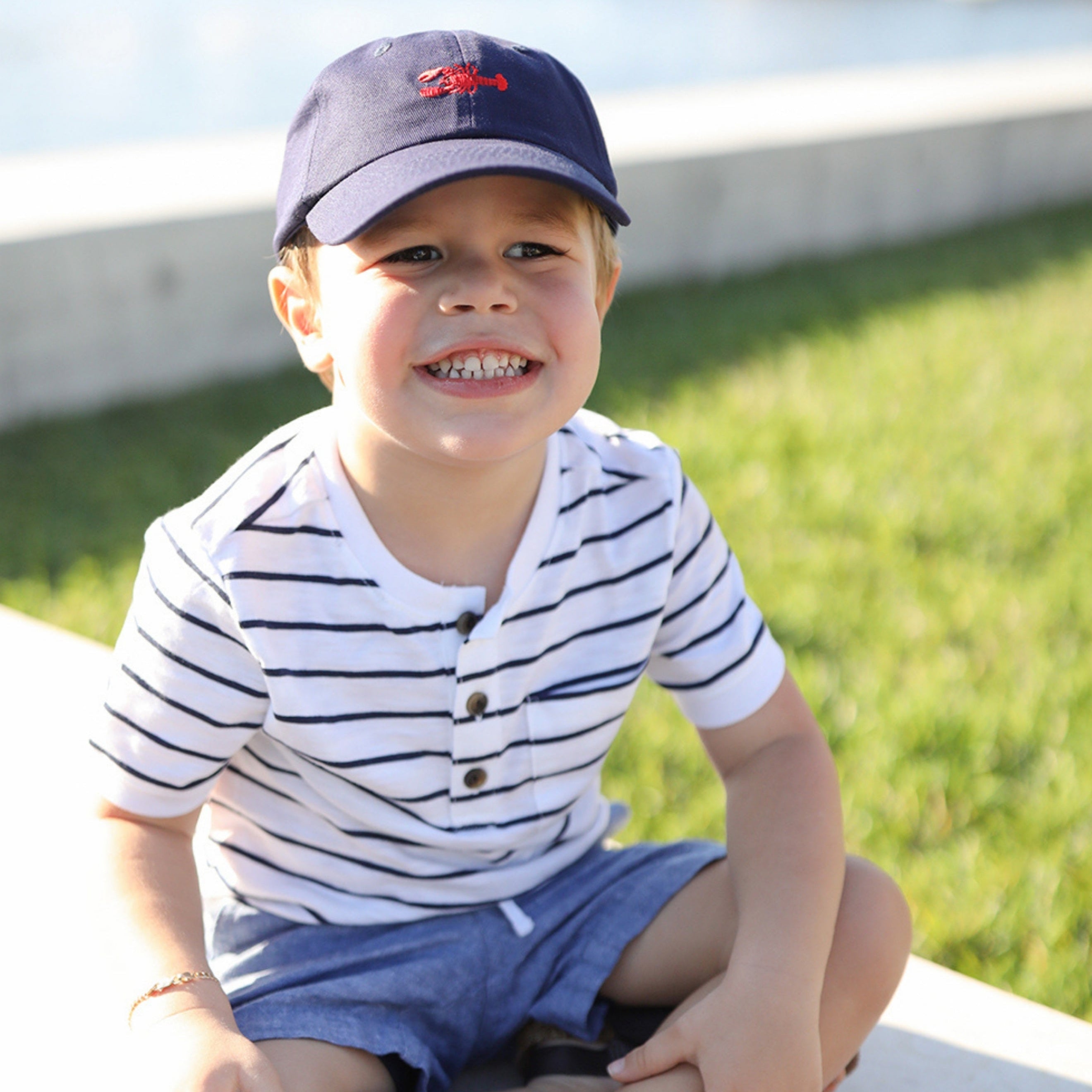 Child wearing a striped shirt and cap sitting on a dock with grass and water in the background