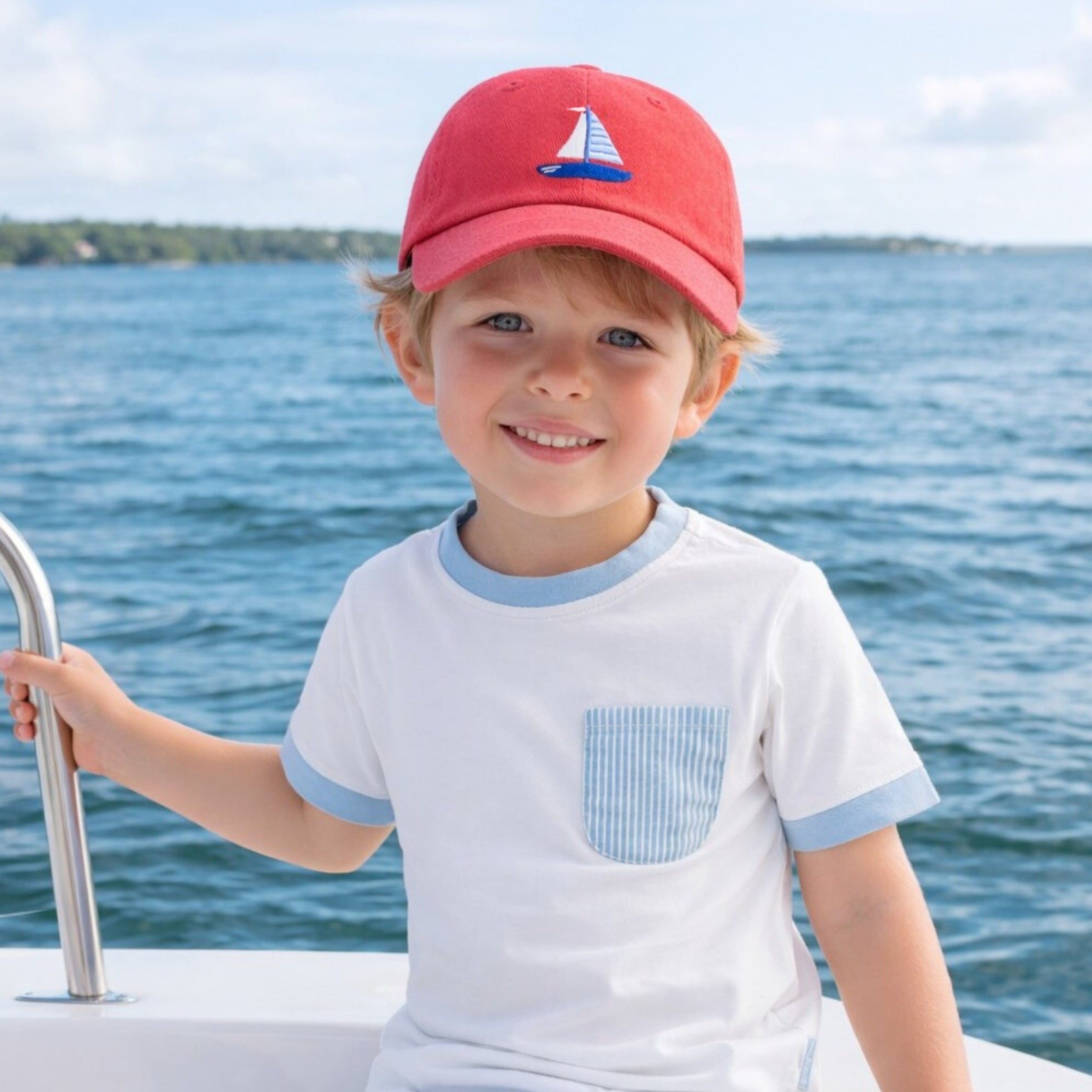 Child wearing a red cap with a sailboat design on a boat with water in the background