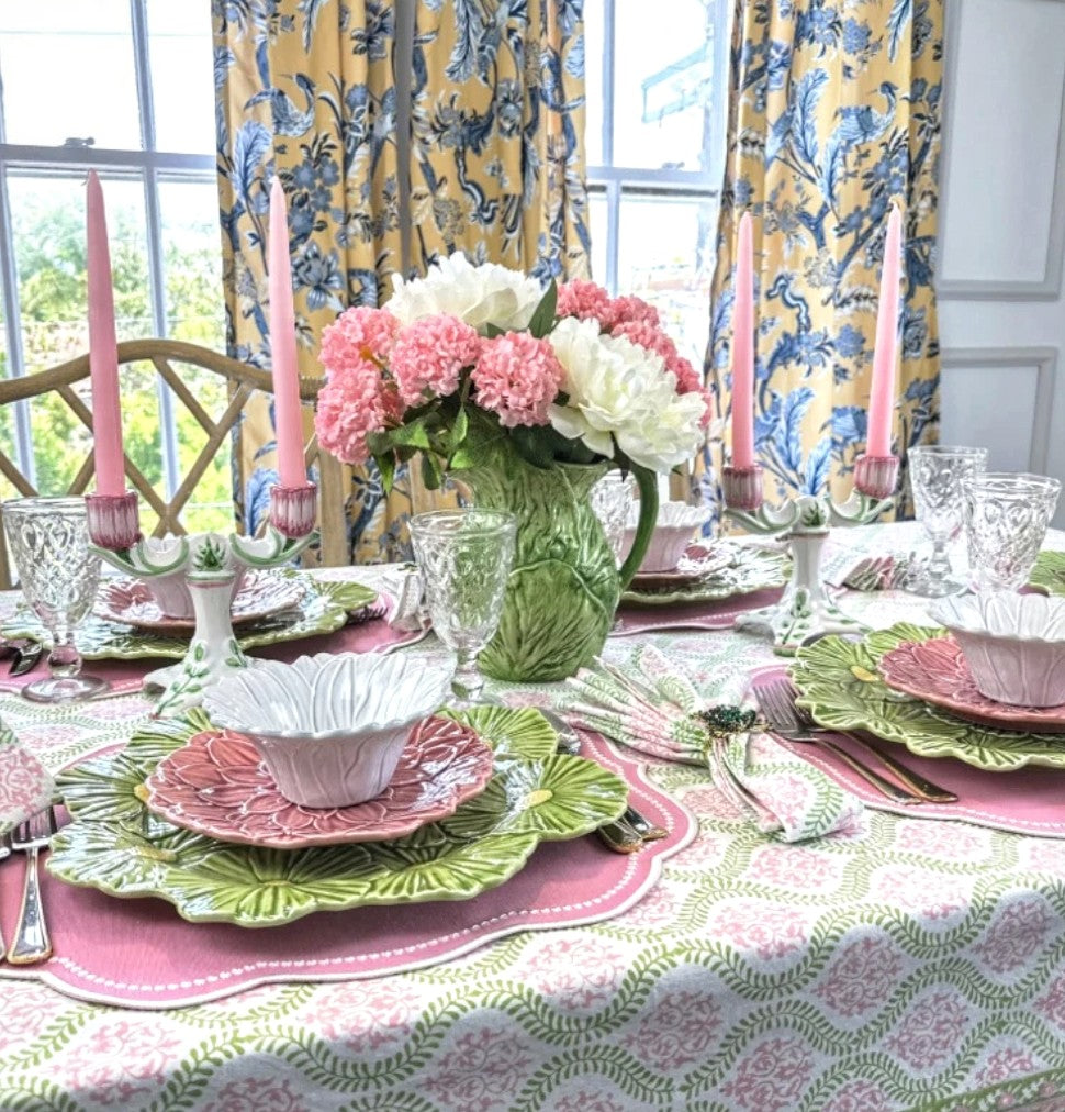 Elegant table setting with pink and green plates, flowers, and candles in a bright room.