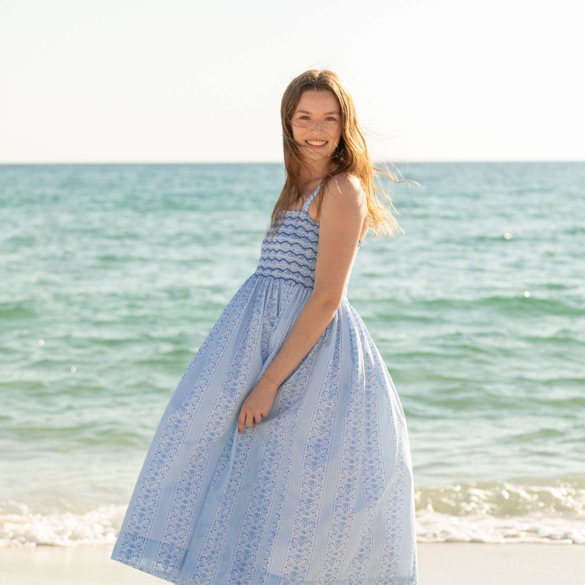 Woman walking along the beach wearing the Henry Duvall Kate Dress, a breezy striped women’s dress with coastal style.