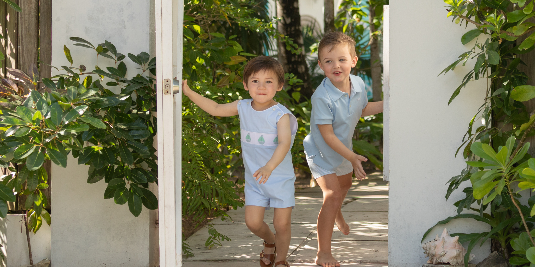 Two children in matching outfits standing in a doorway with greenery around.
