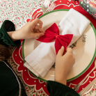 Red velvet bow napkin ties on white napkins, styled on a festive holiday table with holly tablecloth and scalloped placemats – perfect for Christmas entertaining and seasonal celebrations.