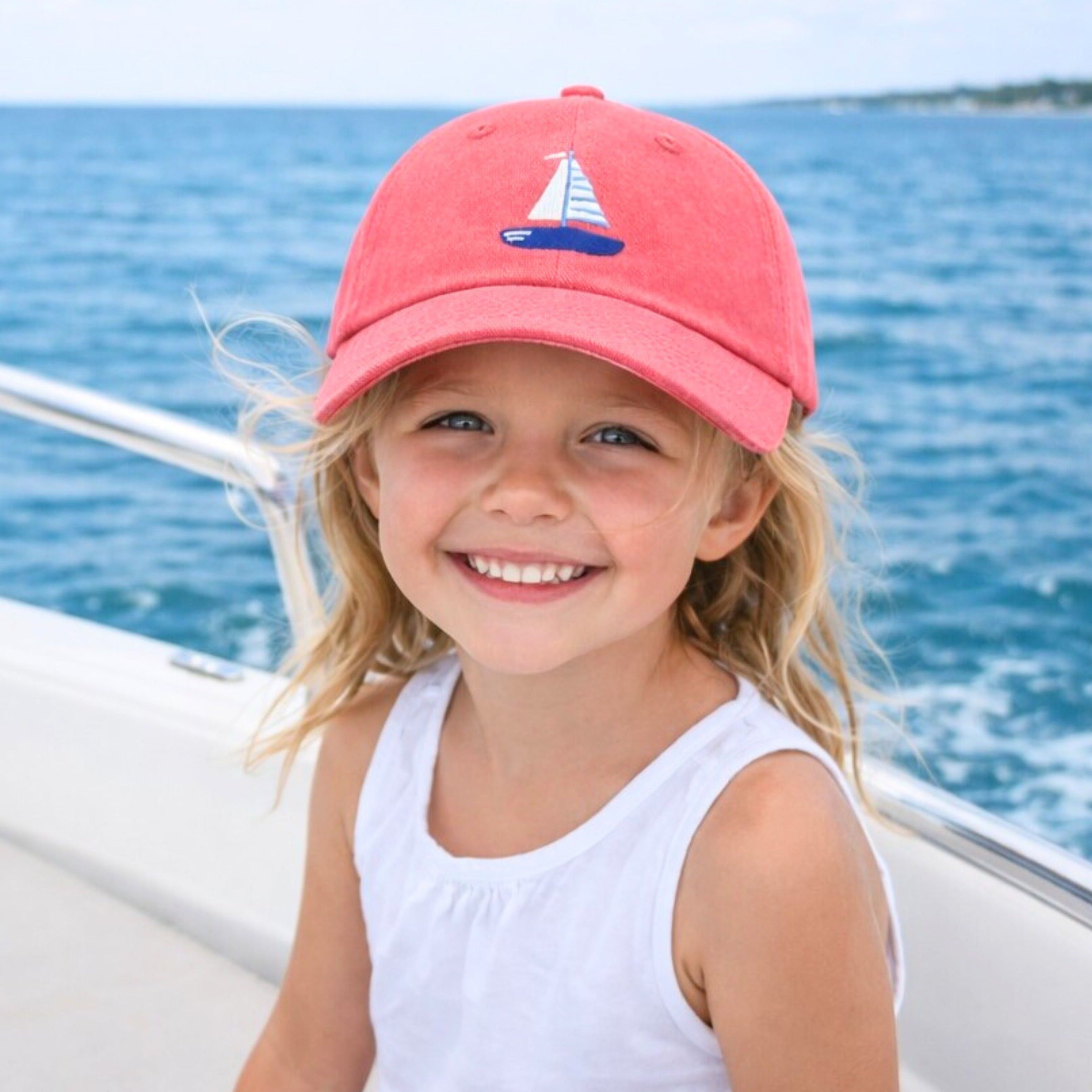 Young girl wearing a pink cap with a sailboat design, sitting on a boat with blue water and sky in the background.