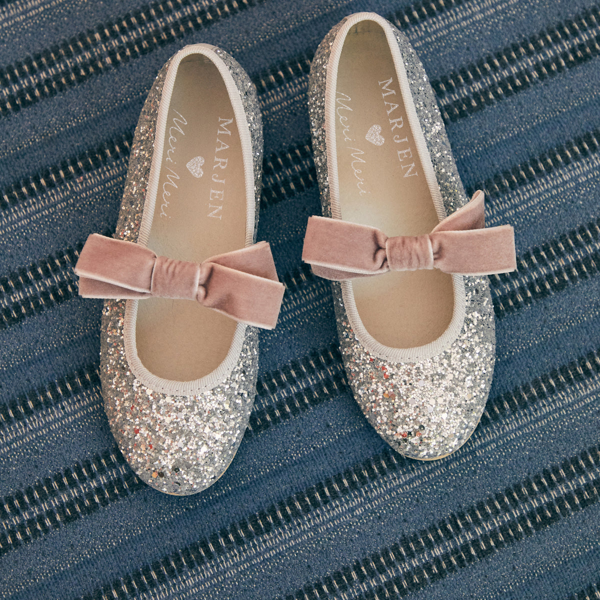 Meri Meri x Marjen Sabrina silver glitter Mary Jane with rose velvet bow, pair on a blue striped rug