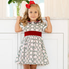 Girls’ holiday dress in Henry Duvall Regent Wreaths red and green Christmas print, featuring a red velvet Peter Pan collar and sash, white pleated trim, puff sleeves, and a full gathered skirt.