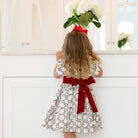 Girls’ holiday dress in Henry Duvall Regent Wreaths red and green Christmas print, featuring a red velvet Peter Pan collar and sash, white pleated trim, puff sleeves, and a full gathered skirt.