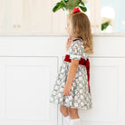 Girls’ holiday dress in Henry Duvall Regent Wreaths red and green Christmas print, featuring a red velvet Peter Pan collar and sash, white pleated trim, puff sleeves, and a full gathered skirt.
