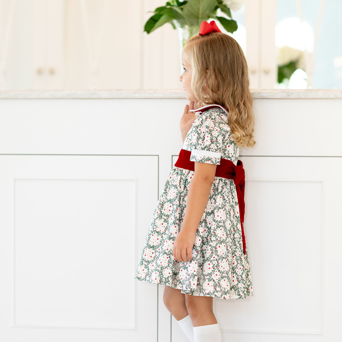 Girls’ holiday dress in Henry Duvall Regent Wreaths red and green Christmas print, featuring a red velvet Peter Pan collar and sash, white pleated trim, puff sleeves, and a full gathered skirt.